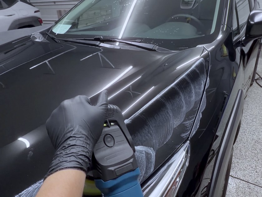 Gloved hands guiding a dual action polisher across glossy black paint, close in.