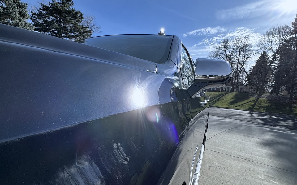 A dark blue Suburban reflecting a treeline and sky along its flank, the reflection running straight and unbroken.